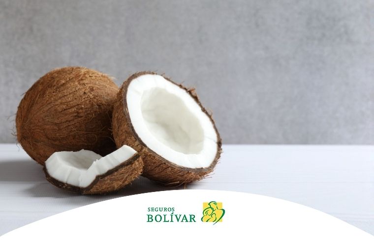 Dos cocos enteros, uno cortado a la mitad revelando la pulpa blanca y un trozo adicional, sobre una superficie clara.