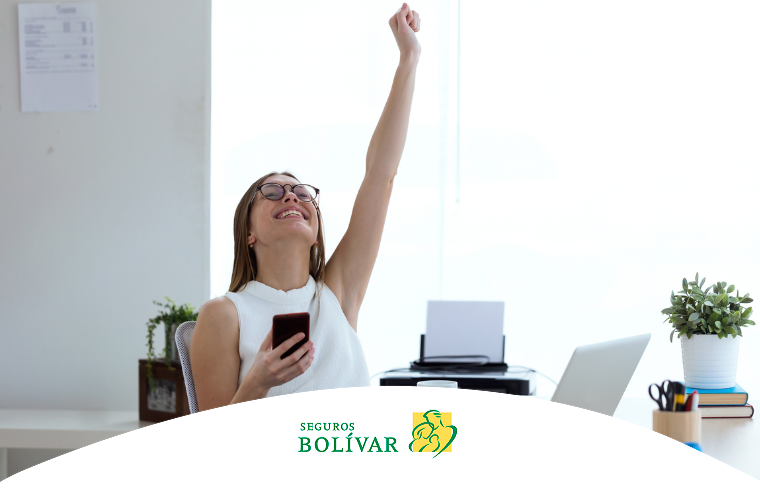 Mujer joven en una oficina, sonriendo con el puño levantado en señal de victoria mientras sostiene un celular, ilustrando éxito o buenas noticias.