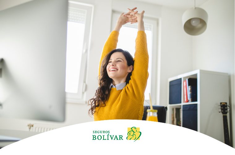 Mujer joven con saco amarillo estirando los brazos frente a su computador, representando pausas activas y bienestar laboral.