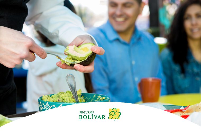 Primer plano de una persona extrayendo la pulpa de un aguacate con una cuchara para preparar guacamole, con una pareja sonriendo al fondo