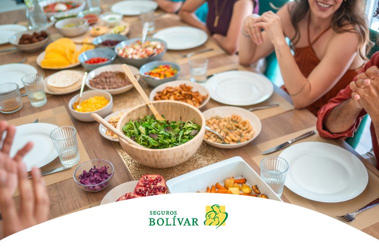 Mesa de madera servida con variedad de platos saludables como ensaladas, granos y frutas, mientras un grupo de personas comparte la comida.