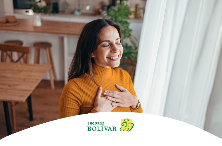 Mujer sonriente con los ojos cerrados y las manos sobre el pecho, junto a una ventana soleada en su cocina, transmitiendo gratitud.